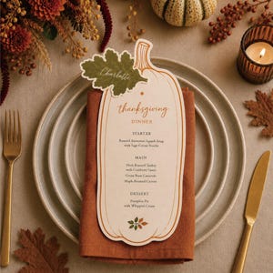 May include: A pumpkin-shaped Thanksgiving dinner menu card with a green leaf name tag reading Charlotte, placed on a rust-colored napkin and stacked ceramic plates. The menu lists courses including Roasted Butternut Squash Soup and Turkey.