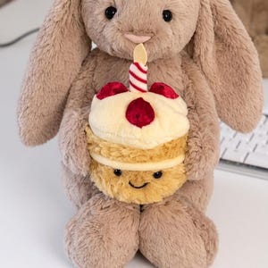 May include: A plush bunny toy in a light brown color, holding a cupcake with a candle. The cupcake has a cream-colored top with red cherry accents and a smiling face on the bottom layer. The candle is red and white striped.