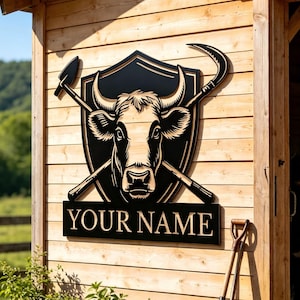 Pode incluir: Uma placa de metal preta com a imagem de uma cabeça de vaca dentro de um escudo, com ferramentas agrícolas cruzadas e o texto "YOUR NAME". A placa está montada numa parede de celeiro de madeira, com plantas em vasos e ferramentas de jardinagem em primeiro plano.