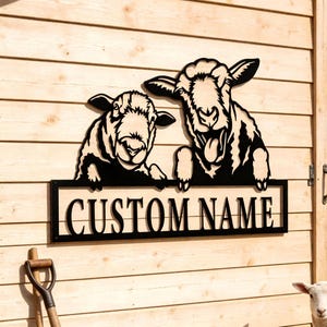 May include: A black metal sign featuring two sheep with the text "CUSTOM NAME" in a rectangular frame. The sign is mounted on a wooden barn wall. A lamb stands in front of the barn.