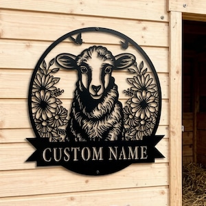 May include: Black metal sheep sign with a floral design and the text "CUSTOM NAME". The sign is mounted on a wooden wall next to a barn. A small white lamb stands in front of the wall.