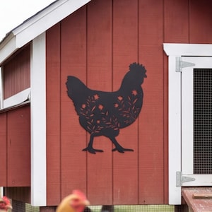 Può includere: Un pezzo di arte murale in metallo nero a forma di gallina con un motivo floreale ritagliato, montato sulla parete esterna di un pollaio in legno rosso. La sagoma presenta intricati disegni di foglie e fiori su tutto il corpo dell'animale.