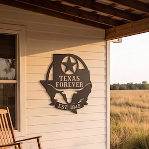 Puede incluir: Un letrero de pared de metal negro con la forma del estado de Texas, que presenta una estrella recortada, la silueta de un buey longhorn y el texto "TEXAS FOREVER" con "EST. 1845" en una pancarta en la parte inferior, sobre una pared blanca.
