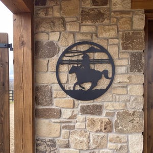 May include: A circular black metal wall art piece featuring a silhouette of a person riding a horse while swinging a lasso. The design includes a horizontal fence line in the background and is mounted on a textured light brown stone wall surface.