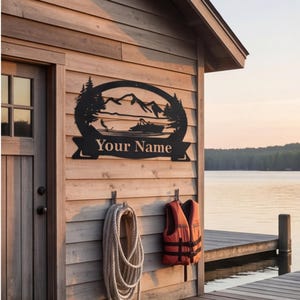 Könnte beinhalten: Ein schwarzes Metall-Wandschild mit einer Silhouette aus Bergen, Tannen und einem Boot auf dem Wasser, mit einem Banner am unteren Rand, das den Text Your Name zeigt. Das Schild ist an der hölzernen Außenwand eines Gebäudes am See befestigt.