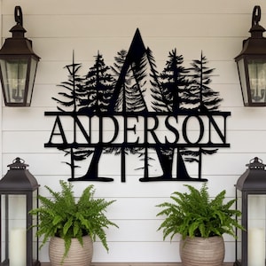 May include: A black metal wall sign featuring a large letter A integrated with a forest of pine trees and the name ANDERSON in capital letters. The sign is displayed on a white horizontal paneled wall between two black lanterns and two ferns.