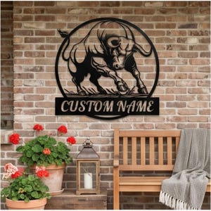 May include: A black metal wall sign featuring a circular frame with a silhouette of a charging bull. Below the bull, a rectangular banner contains the text CUSTOM NAME in bold, capital letters. The sign is mounted on a textured brick wall.