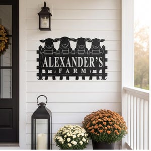 Puede incluir: Un letrero de pared de metal negro con un diseño recortado de cuatro ovejas asomándose sobre una cerca. El texto "ALEXANDER'S FARM" se muestra de forma destacada en el centro. La pieza tiene un acabado mate y una forma rectangular.