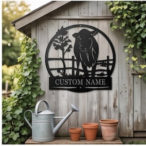 May include: A circular black metal wall sign featuring a silhouette of a cow standing in front of a fence and a tree. The sign includes a rectangular base section with the text CUSTOM NAME centered in capital letters in a serif font.