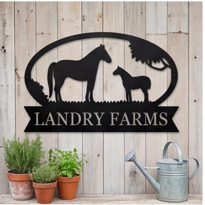 May include: A black metal oval sign featuring a silhouette of a horse and a foal standing in grass under a tree. Below the oval, a black banner displays the text LANDRY FARMS in white capital letters. The sign has a flat, matte finish.