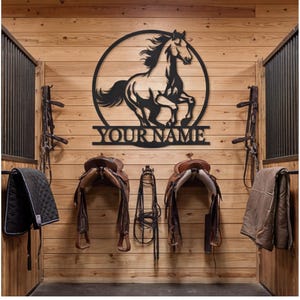 May include: A black metal wall sign featuring a circular frame with a silhouette of a galloping horse and the text YOUR NAME centered at the bottom. The sign is mounted on a light-colored wooden wall above two brown leather horse saddles.