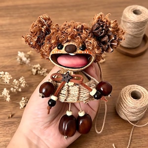 May include: A handcrafted doll with brown curly hair, a smiling face, and a pink tongue. The doll's body is made of woven material, decorated with brown beads and acorn-shaped elements. The doll is suspended by a string.