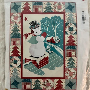 以下が含まれることがあります： WINTER NIGHT IN SNOWTOWNというタイトルのキルトキットパッケージ。煙突の上にシルクハットをかぶった雪だるまが描かれた中央のイラストと、赤と青緑色の木、家、星のパッチワーク模様の正方形で構成された縁取りが特徴です。