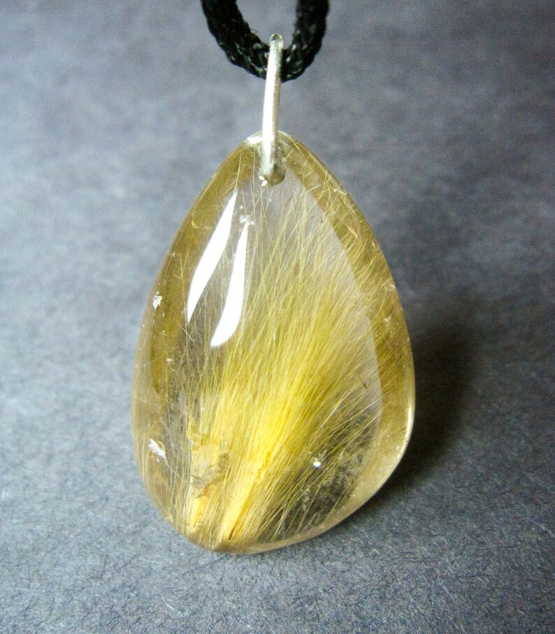 May include: A teardrop-shaped pendant with a clear quartz crystal containing a golden-brown hair-like inclusion. The pendant is attached to a black cord.