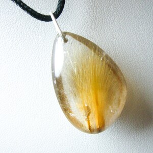 May include: A teardrop-shaped pendant necklace with a clear quartz crystal containing a golden brown feather. The pendant is attached to a black cord.