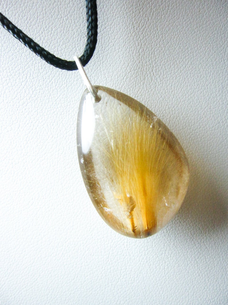 May include: A teardrop-shaped pendant necklace with a clear quartz crystal containing a golden brown feather. The pendant is attached to a black cord.