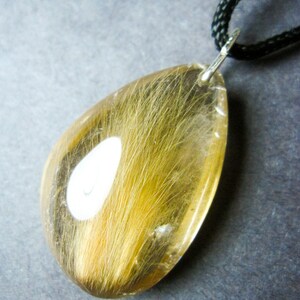May include: A teardrop-shaped pendant with a clear resin filled with a strand of light brown hair. The pendant is attached to a black cord.