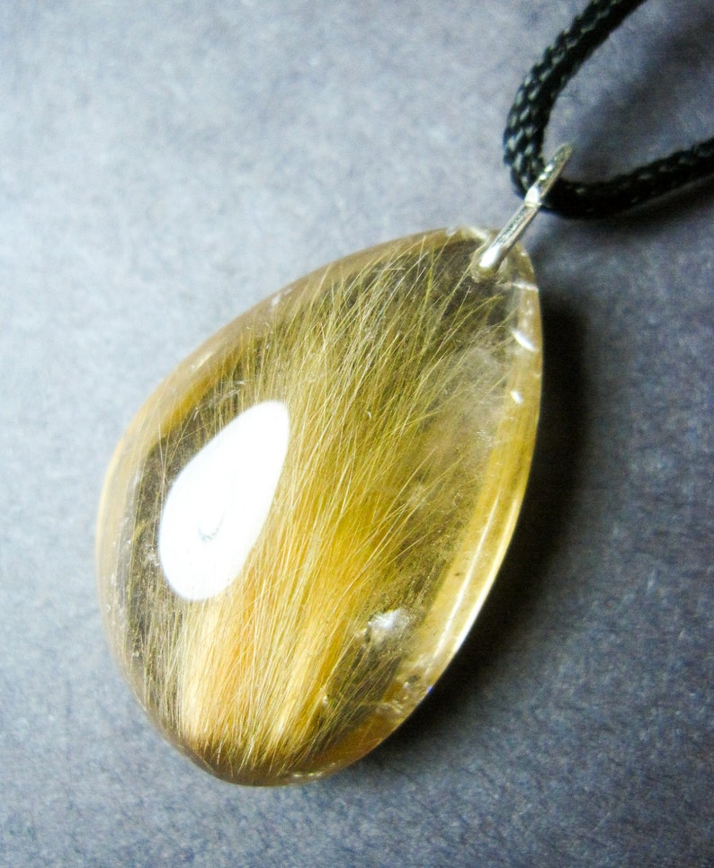 May include: A teardrop-shaped pendant with a clear resin filled with a strand of light brown hair. The pendant is attached to a black cord.