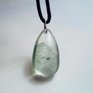 May include: A clear, teardrop-shaped pendant with a silver bail hangs from a black cord. The pendant has a light green hue and contains small, textured inclusions. The background is a plain white surface.