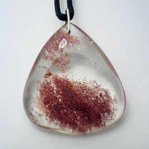 May include: A clear, triangular pendant with a reddish-brown inclusion, suspended from a black cord. The pendant is made of a transparent material, showcasing the natural, organic patterns within. The cord is attached with a silver-colored bail.