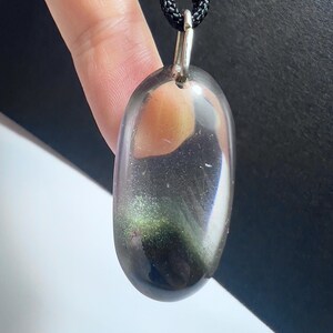 May include: An oval-shaped pendant with a silver bail, suspended from a black cord. The pendant is a translucent, iridescent stone, displaying shades of green, gold, and purple. The stone has a polished finish, reflecting light.