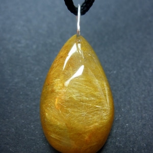 May include: A gold teardrop-shaped pendant with a black cord. The pendant has a translucent, golden color with visible gold flecks.