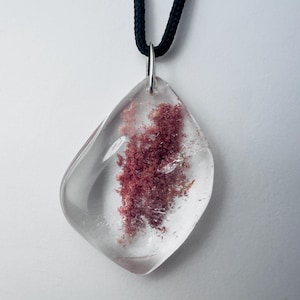 May include: A clear, irregularly shaped pendant with a reddish-brown inclusion, suspended from a black cord. The pendant is attached to a silver-colored bail. The background is a plain white.