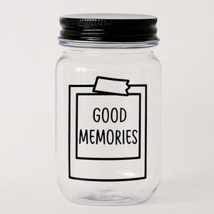 May include: A clear glass jar with a black lid. The jar has a white rectangular label with the words "GOOD MEMORIES" in black. The label is taped to the jar with a piece of tape. The jar is empty.