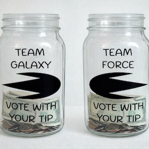 May include: Two clear glass jars with lids, each filled with money. One jar reads "TEAM GALAXY" and the other reads "TEAM FORCE." Both jars have a black graphic and the text "VOTE WITH YOUR TIP."