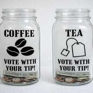 May include: Two clear glass jars labeled "COFFEE" and "TEA" with black text and illustrations. The coffee jar has coffee bean graphics, and the tea jar has a tea bag graphic. Both jars read "VOTE WITH YOUR TIP!" and contain money.