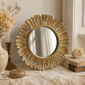 May include: A round mirror with a gold-toned frame featuring a decorative border of sculpted feathers arranged in a sunburst pattern. The reflective glass surface is circular and set within the center of the detailed, textured metallic frame.
