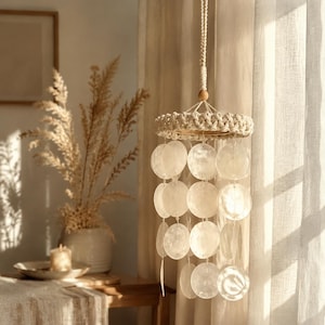May include: A hanging wind chime with a macrame top and a series of white, round shell discs. The chime is suspended from a natural rope and wooden bead. The decor is in a neutral color palette.