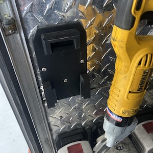 May include: A black plastic tool holder mounted on a diamond-plate metal surface. A yellow and black power tool is partially visible, along with several battery packs. The holder is secured with screws.
