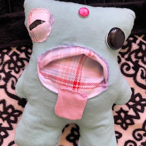 Puede incluir: Un juguete de peluche hecho a mano, de color azul claro, con detalles cosidos. Cuenta con un botón rosa, un botón negro y un ojo cosido. El juguete tiene una barriga abierta que revela un patrón de cuadros. Un pequeño rectángulo rosa está cosido debajo de la barriga.