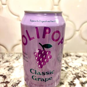 May include: A purple can of Olipop Classic Grape soda. The can features the brand name in large, pink letters, a grape illustration, and the text "Supports Digestive Health." The can contains 12 fl oz (355 ml) of soda.