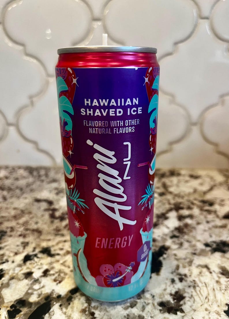 May include: A cylindrical candle in a can, designed to resemble a can of Alani Nu Hawaiian Shaved Ice energy drink. The can is primarily red and purple, with teal and white accents, and features the text "Hawaiian Shaved Ice" and "Energy".