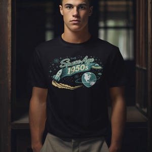 May include: Black t-shirt with a retro space-themed graphic. The design features a rocket, a planet, and a flying saucer, with the text "The Space Age 1950s" in a teal and white color scheme. The shirt is worn by a person.