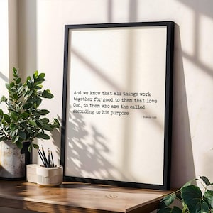 Puede incluir: Una impresión enmarcada con un borde negro muestra el texto "Y sabemos que a los que aman a Dios, todas las cosas les ayudan a bien, esto es, a los que conforme a su propósito son llamados - Romanos 8:28." Una planta en maceta y un portalápices están sobre una superficie de madera.