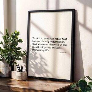 May include: A framed print with the text "For God so loved the world..." in black font. The print is in a black frame and sits on a wooden surface next to a potted plant and a pencil holder.
