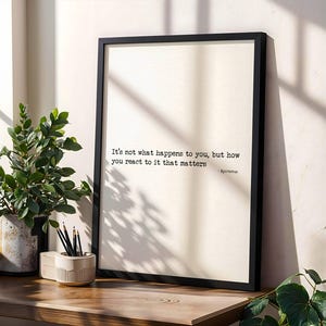 May include: A framed print with a black border displays the quote, "It's not what happens to you, but how you react to it that matters." A small ceramic pencil holder and a potted plant sit on a wooden surface.