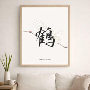 以下が含まれることがあります： 鶴が飛んでいる様子を描いた額入りのアートプリント。日本語の書道と英語のテキスト「Tsuru - Crane」が特徴です。作品は、クリーム色の背景と天然木のフレームを使用したミニマリストデザインです。