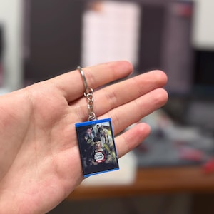 Peut inclure: Un porte-clés rectangulaire avec une image colorée de personnages de la série animée "Demon Slayer". Le porte-clés a un bord bleu et un porte-clés argenté. Le texte "Demon Slayer" est visible sur l'image.