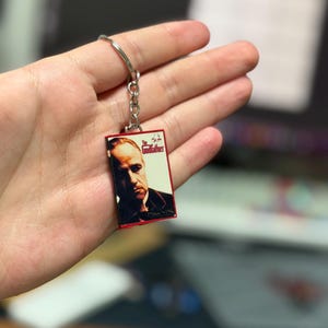 May include: A rectangular keychain featuring a portrait of Marlon Brando from "The Godfather" movie. The keychain has a red border and a silver keyring. The movie title "The Godfather" is printed on the keychain.
