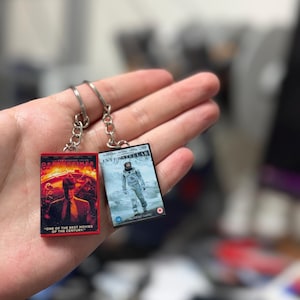 May include: Two movie-themed keychains. One keychain features the movie "Oppenheimer" with a red and black design and the text "ONE OF THE BEST MOVIES OF THE CENTURY." The other keychain is for the movie "Interstellar."