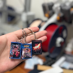 Op de afbeelding: Drie miniatuur films DVD sleutelhangers. De sleutelhangers tonen de covers van "Doctor Strange in the Multiverse of Madness", "Avengers" en "The Dark Knight Rises". Elke sleutelhanger heeft een zilveren sleutelring.