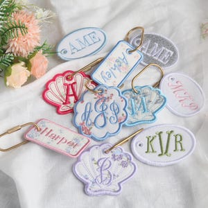 May include: A collection of embroidered keychains in various shapes including ovals, rectangles, and seashells. The keychains feature personalised names and monograms in blue, pink, and green thread, attached to gold-toned metal rings.