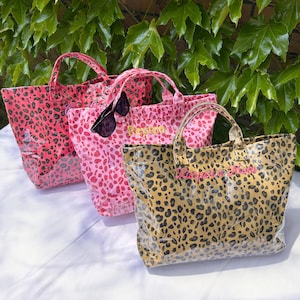 Peut inclure: Trois sacs fourre-tout à imprimé léopard, rouge, rose et beige. Les sacs ont des poignées et sont personnalisés avec des noms en lettres dorées et roses. Des lunettes de soleil sont posées sur l'un des sacs.