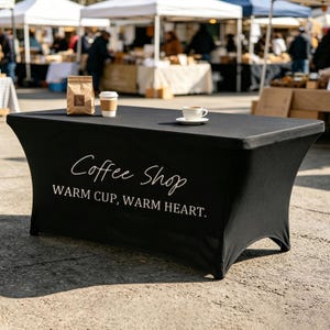 May include: A black fitted table cover with white script reading "Coffee Shop, Warm Cup, Warm Heart". A coffee cup, a paper cup, and a bag of coffee beans are on the table. The setting is an outdoor market with stalls in the background.