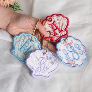 May include: Four embroidered shell-shaped keychains with gold-toned metal carabiner clips. Each shell features a unique floral pattern and a prominent embroidered monogram letter in red, blue, or purple thread, including the letters A, M, B, and AG.