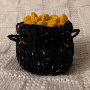 May include: A black crocheted pot filled with yellow crocheted gold coins. The pot has a speckled pattern and a drawstring closure.